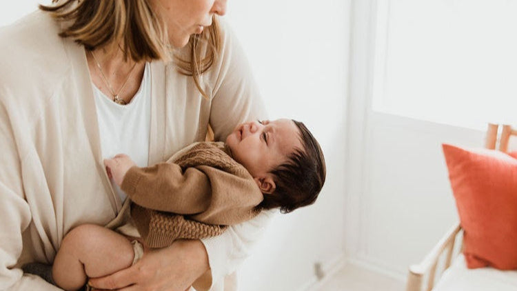 Woman holding a baby in a cozy indoor setting with a chair and cushions.